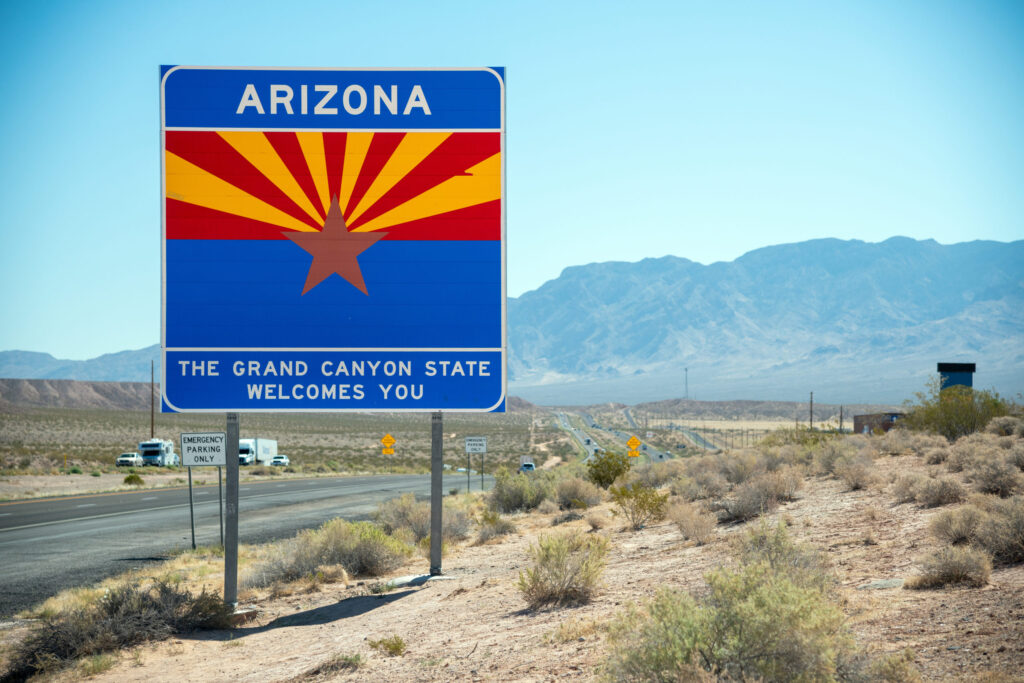 Arizona state line sign reading The Grand Canyon State Welcomes You beside a desert highway, with trucks on the road and mountains behind