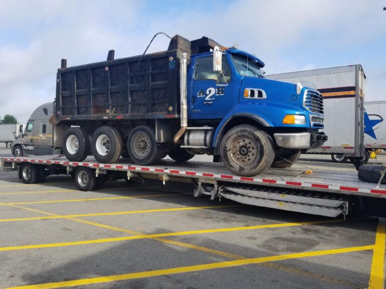 Blue tri-axle dump truck loaded on a trailer deck in a truck lot