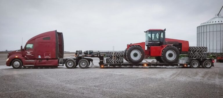 Red four-wheel-drive farm tractor in the low well of an RGN trailer behind a maroon semi, beside a grain bin