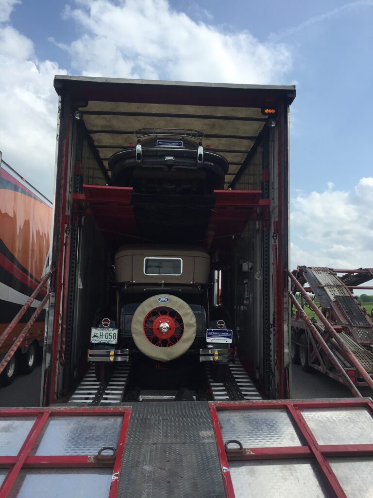 Classic car with a rear-mounted spare tire strapped on the lower deck inside an enclosed two-level car trailer